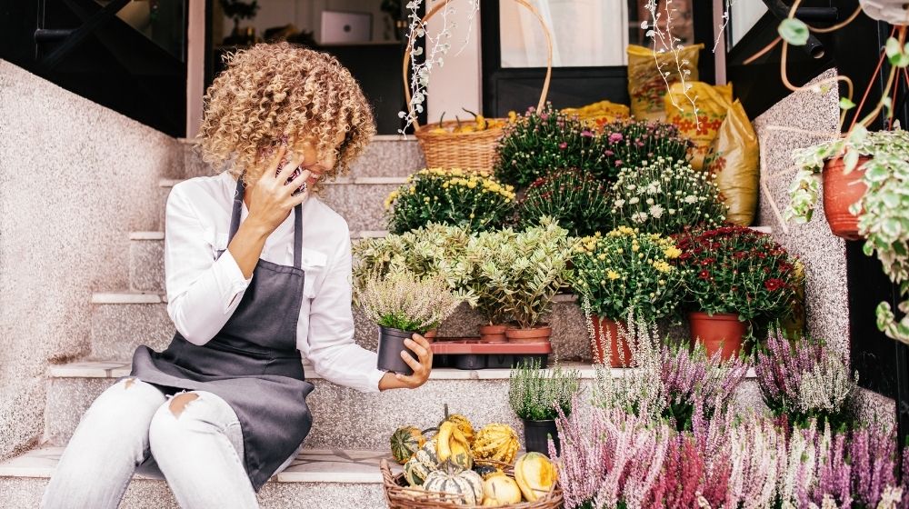 Woman sitting infront of a flower shop while talking on her phone and holding a plant | FE | 3 Franchise Marketing Ideas to Boost Local Business