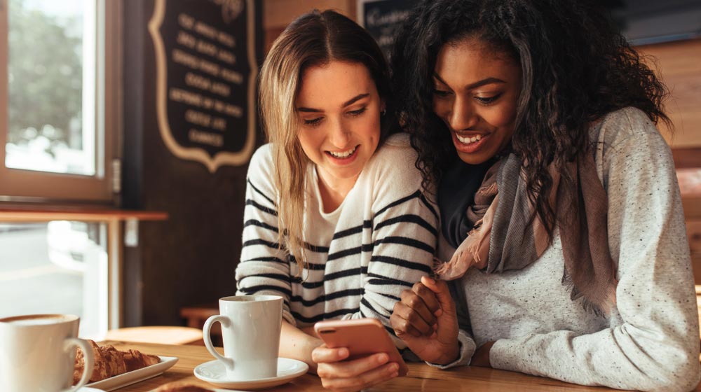 two-young-women-caucasian-black-scrolling-on-phone-while-having-breakfast-in-coffee-shop | feature | Convert Searches to Sales with LOCL’s Google Post Feature